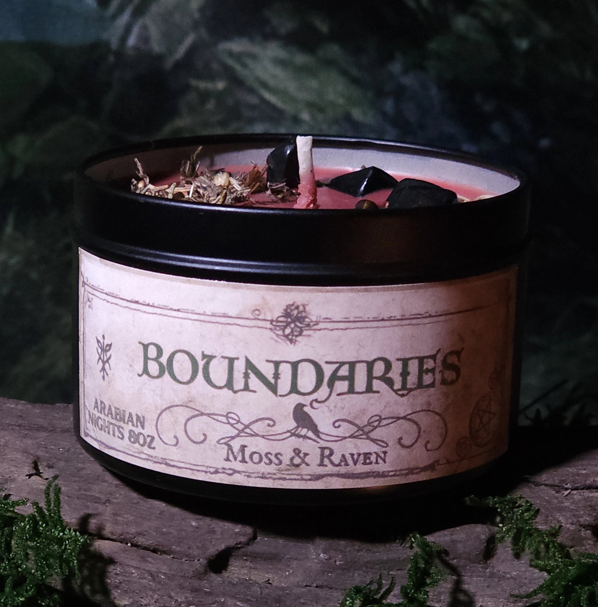 Candle in a container labeled 'Boundaries' by Moss & Raven on a dark background with greenery.