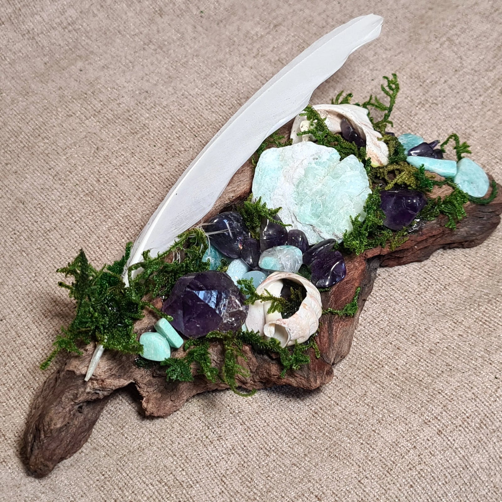Driftwood altar with Amazonite, Amethyst, and moss beneath a white feather – handmade by Moss & Raven.
