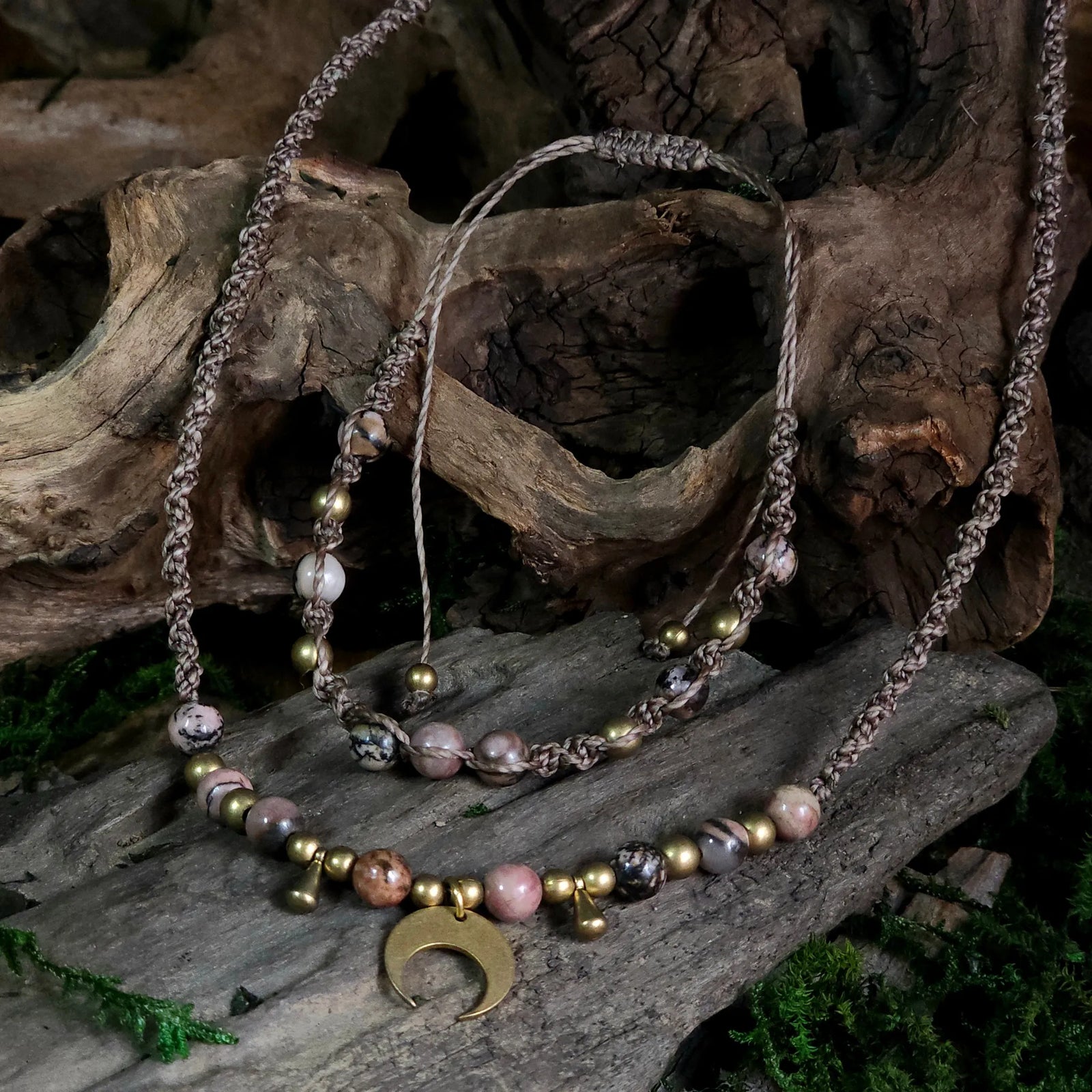 Braided cord necklace and bracelet set with rhodonite beads, brass accents, and a brass crescent moon pendant, displayed on driftwood with natural moss.