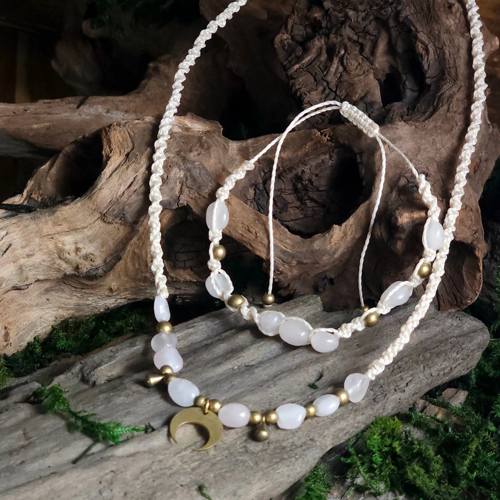 Handwoven ivory macramé necklace and bracelet set with rose quartz beads, brass accents, and a brass crescent moon pendant, displayed on driftwood and moss.